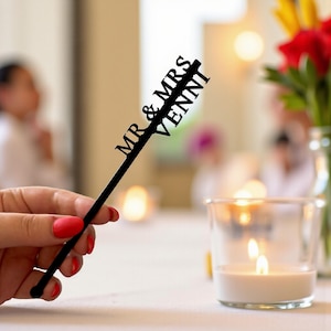 May include: A black cake topper with the words "MR & MRS VENNI" cut out in a script font. The topper is held by a person with red nail polish. A lit candle and flowers are in the background.