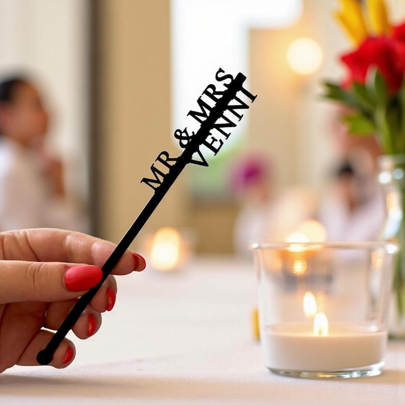 May include: A black cake topper with the words "MR & MRS VENNI" cut out in a script font. The topper is held by a person with red nail polish. A lit candle and flowers are in the background.