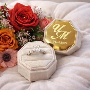 Könnte beinhalten: Ein Diamant-Verlobungsring in einer hellgrauen Samt-Ringbox, neben einer passenden Box mit goldfarbenen Buchstaben und dem Datum 04.04.2025. Ein Strauß aus roten und orangen Rosen und anderen Blumen befindet sich im Hintergrund.