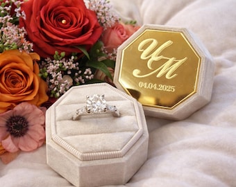 May include: A diamond engagement ring in a light gray velvet ring box, next to a matching box with gold lettering and the date 04.04.2025. A bouquet of red and orange roses and other flowers is in the background.