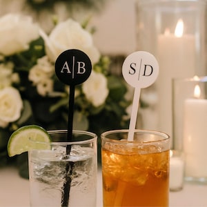 May include: Two cocktails with personalized drink stirrers. One stirrer is black with "AB" in white, the other is white with "SD" in black. One drink has a lime wedge, the other has ice.