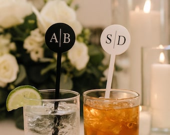 May include: Two cocktails with personalized drink stirrers. One stirrer is black with "AB" in white, the other is white with "SD" in black. One drink has a lime wedge, the other has ice.