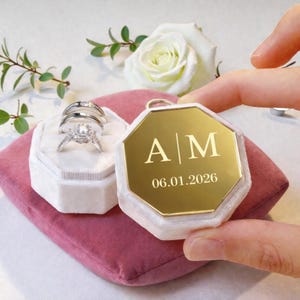 May include: A close-up of a wedding ring set in a white velvet box, next to a gold-colored, octagonal keepsake box with the initials "A | M" and the date "06.01.2026" engraved on it. The boxes rest on a pink velvet surface.