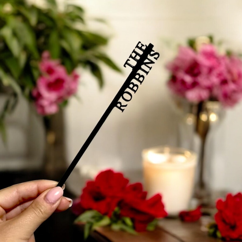 May include: A black cake topper with the text "THE ROBBINS" in a vertical arrangement. The topper is held in front of a blurred background with flowers and a candle.