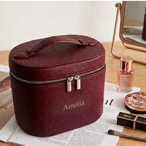 May include: A deep burgundy, oval-shaped cosmetic case with a textured finish and a top handle. The front features the name Amelia in gold lettering. A silver-toned double zipper closure secures the main compartment of this structured travel bag.