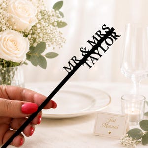 May include: A black cake topper featuring the text MR & MRS TAYLOR in a bold, sans-serif font. The topper consists of a long, thin, straight vertical stem that extends upward into the personalized lettering, creating a clean and modern aesthetic.