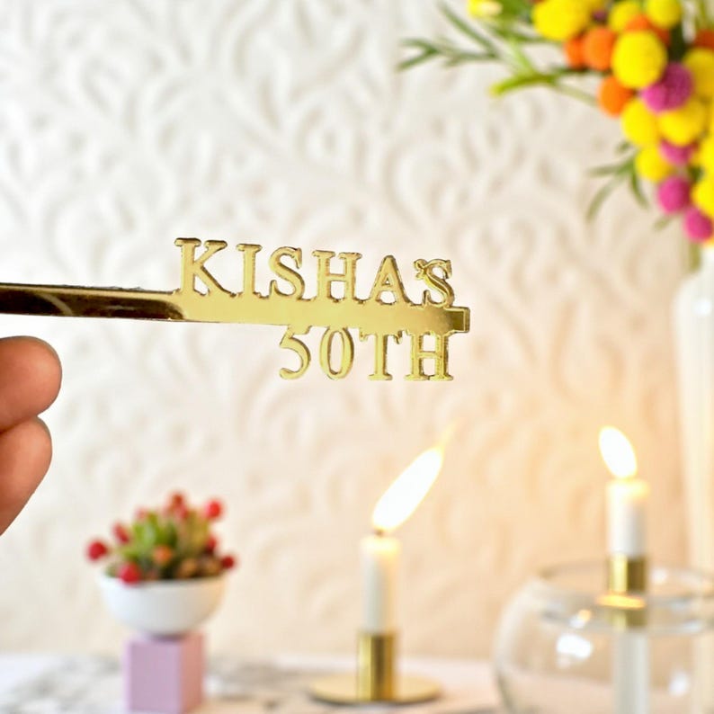May include: A gold-colored cake topper with the name "KISHAS" and the number "50TH" cut out. The topper is held in front of a blurred background with lit candles and flowers, suggesting a birthday celebration.