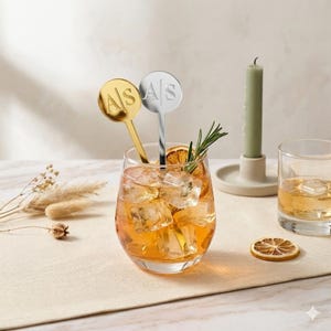 May include: Two round metal cocktail stirrers, one gold and one silver, each featuring the engraved letters A|S. The stirrers are placed inside a glass filled with an amber-colored liquid, ice cubes, and a sprig of rosemary, alongside a candle.