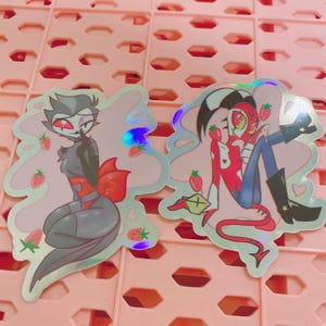 May include: Two holographic stickers featuring cartoon characters with strawberry accents. One sticker depicts a character in a black and gray outfit with a red bow. The other shows two characters embracing, with red and blue clothing.