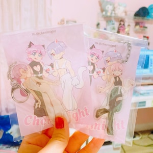 May include: Two clear acrylic charms featuring anime-style characters with cat ears. One charm has a keychain attached. The charms are on pink backing cards with the text "Cheri Might" and the Instagram handle "@cherimight".