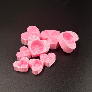 May include: A collection of small, light pink heart-shaped objects. Each heart features a recessed circular center with a radiating sunburst pattern etched into the surface. The items are arranged in a cluster, displaying a smooth, matte finish.