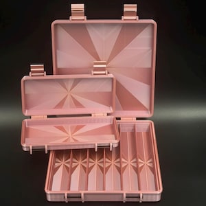 May include: A set of three rose gold storage cases with hinged lids. The cases feature a geometric sunburst pattern on the interior surfaces. The largest case is square, the medium case is rectangular, and the smallest case is a slim rectangle.