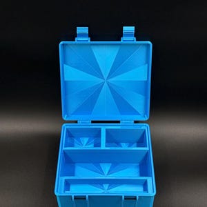 May include: A bright blue plastic storage case with an open hinged lid. The interior features a base divided into four distinct compartments of varying sizes. Both the lid and the base display a subtle, radiating sunburst pattern texture.