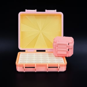 May include: A pink, open case with a yellow interior and a smaller, matching pink case. The open case has a tray with multiple circular compartments. The cases are on a black background.