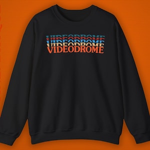 May include: Black crewneck sweatshirt with the word "VIDEODROME" stacked in a retro font. The text is in shades of blue, red, and orange. The background is a textured orange.