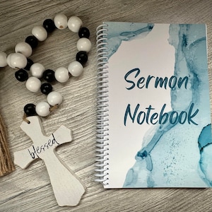 Peut inclure: Un carnet blanc avec un motif aquarelle bleu et le texte "Sermon Notebook". On voit &eacute;galement une croix en bois avec le mot "blessed" &eacute;crit dessus et une guirlande de perles noires et blanches avec un pompon brun.