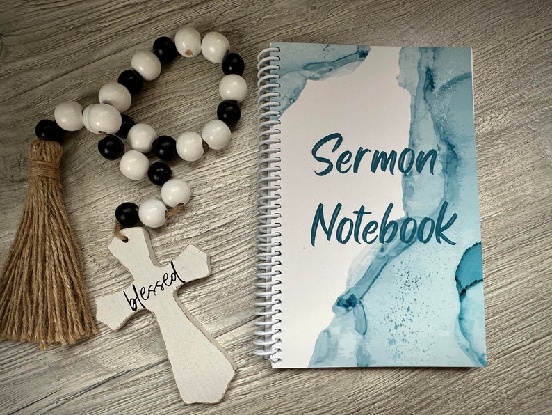 Peut inclure: Un carnet blanc avec un motif aquarelle bleu et le texte "Sermon Notebook". On voit &eacute;galement une croix en bois avec le mot "blessed" &eacute;crit dessus et une guirlande de perles noires et blanches avec un pompon brun.