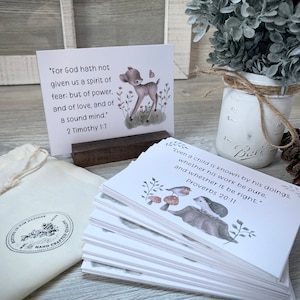 May include: A set of 20 white cards with inspirational bible verses and cute animal illustrations. The cards are stacked on a wooden table with a small white bag and a mason jar with a sprig of greenery. The text on the top card reads "For God hath not given us a spirit of fear: but of power, and of love, and of a sound mind." 2 Timothy 1:7. The text on the bottom card reads "Even a child is known by his doings, whether his work be pure, and whether it be right." Proverbs 20:11.