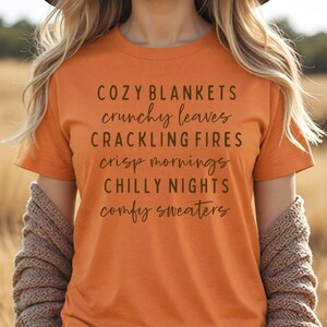 May include: An orange short-sleeved t-shirt featuring dark brown text that reads: COZY BLANKETS, crunchy leaves, CRACKLING FIRES, crisp mornings, CHILLY NIGHTS, comfy sweaters. The shirt has a crew neckline and a standard fit for casual wear.