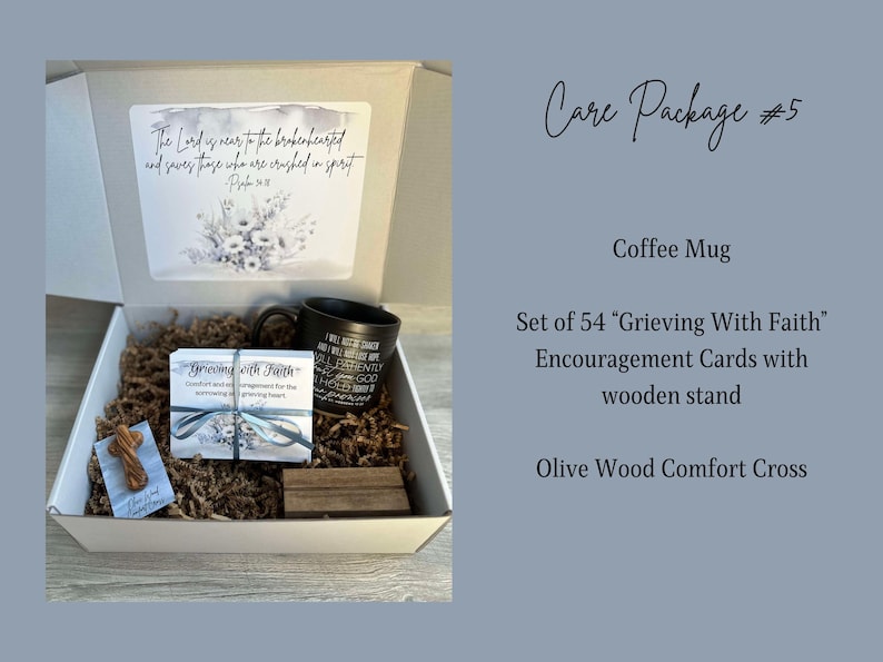 May include: A white gift box filled with brown paper. The box contains a black coffee mug, a set of 54 "Grieving With Faith" encouragement cards with a wooden stand, and a small olive wood comfort cross. The box lid has a card with a floral design and the text "The Lord is near to the brokenhearted and saves those who are crushed in spirit - Psalm 34:18"