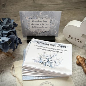 May include: A set of cards with a blue and white floral design. The cards have the text "Grieving with Faith" and "Comfort and encouragement for the sorrowing and grieving heart." The cards are stacked on a wooden surface next to a white heart with the word "Faith" written on it. There is a small wooden cross and a blue hydrangea in the background.