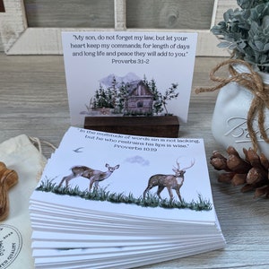 May include: A stack of white cards with inspirational quotes from the book of Proverbs. Each card features a watercolor illustration of a cabin in the woods, a deer, or a bird. The text on the cards reads "My son, do not forget my law, but let your heart keep my commands; for length of days and long life and peace they will add to you." Proverbs 3:1-2 and "In the multitude of words sin is not lacking, but he who restrains his lips is wise." Proverbs 10:19