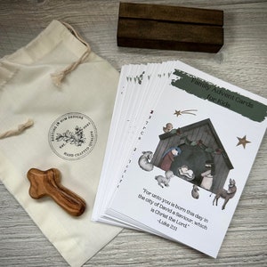 May include: A set of 24 white cards with a green title bar that reads "Family Advent Cards for Kids". The cards feature a nativity scene illustration with a quote from Luke 2:11. The cards are packaged in a white muslin bag with a brown wooden cross and a brown wooden block.