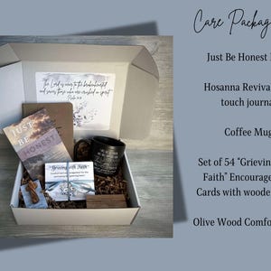 May include: A white gift box filled with a care package. The box contains a book titled "Just Be Honest", a soft touch journal, a coffee mug, a set of 54 encouragement cards with a wooden stand, and an olive wood comfort cross.