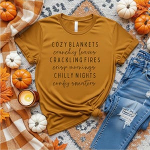 May include: A mustard yellow t-shirt featuring the black text: COZY BLANKETS, crunchy leaves, CRACKLING FIRES, crisp mornings, CHILLY NIGHTS, comfy sweaters. The shirt is surrounded by small orange and white pumpkins, a plaid blanket, and blue jeans.