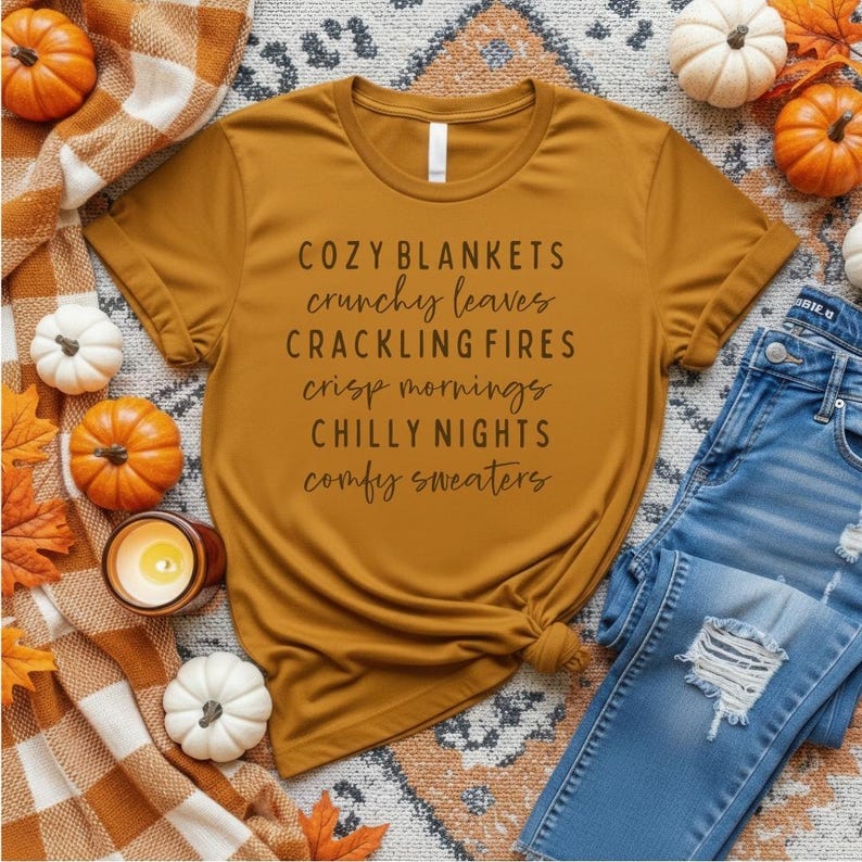May include: A mustard yellow t-shirt featuring the black text: COZY BLANKETS, crunchy leaves, CRACKLING FIRES, crisp mornings, CHILLY NIGHTS, comfy sweaters. The shirt is surrounded by small orange and white pumpkins, a plaid blanket, and blue jeans.