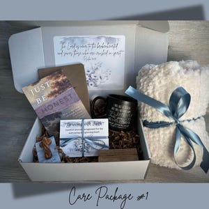 May include: A white gift box with a blue ribbon and a white blanket. The box contains a book titled "Just Be Honest", a mug with a quote about faith, a wooden cross, and a card that says "Grieving with Faith".