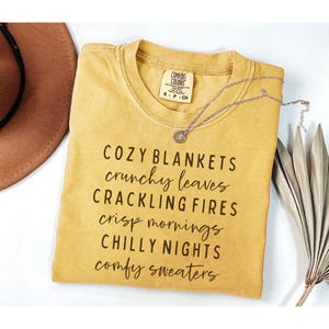May include: A mustard yellow short-sleeve t-shirt with a crew neckline. The shirt features black text that reads: COZY BLANKETS, crunchy leaves, CRACKLING FIRES, crisp mornings, CHILLY NIGHTS, comfy sweaters. A thin silver chain necklace rests on top.