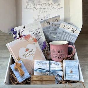 May include: A white gift box filled with items for grieving a loved one. The box contains a book titled "Loved Baby", a mug that says "Be Still & Know That I Am God", a wooden cross, a small card that says "Grieving with Faith", and a necklace with a heart charm. The box is filled with shredded paper.
