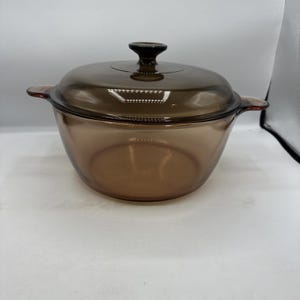 May include: A brown glass casserole dish with a matching lid. The dish has two side handles and a dark knob on the lid. The glass is transparent, allowing visibility of the contents. The dish is designed for cooking and serving.