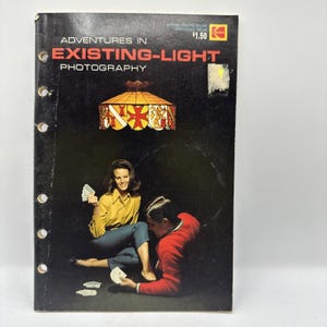 May include: A vintage photography book titled "Adventures in Existing-Light Photography" with a black cover. The cover features a couple playing cards under a decorative lamp. The book has a Kodak logo and a price tag.