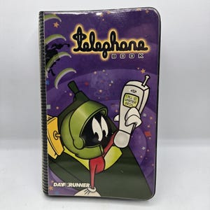 May include: A purple Day Runner telephone book featuring a graphic of Marvin the Martian holding a white mobile phone. The screen displays the text CALL ID: WARMER BROS. The cover has a black textured spine and a glossy finish with stars.