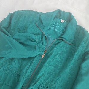 May include: A teal zip-up jacket featuring a tonal floral jacquard pattern, a pointed collar, and elasticized cuffs. The interior label displays the text FU DA International. The garment includes a full-length metal zipper closure and two front pockets.