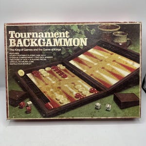 May include: Vintage "Tournament Backgammon" game set in a box. The box features the game's name and features. Inside, the game board is open, displaying red and white playing pieces, dice, and dice shakers. The game set is designed for two players.