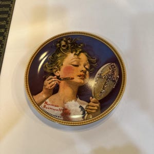 May include: Decorative plate featuring a young person applying lipstick, holding a mirror. The plate has a gold rim and a blue background. The plate is signed "Norman Rockwell".