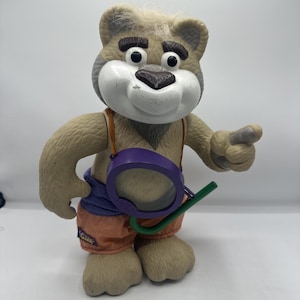 May include: A vintage, tan-colored bear figurine with a white face, wearing orange and purple shorts and a purple accessory. The bear is pointing with its right hand and has a green accessory attached to the purple accessory.