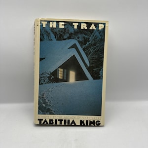 May include: A paperback book titled "The Trap" by Tabitha King. The cover features a snow-covered cabin with a lit window against a backdrop of a snowy forest. The book's title is in white block letters.