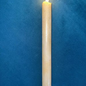 May include: A lit, cream-colored taper candle with a flickering flame. The candle has a textured surface and a white base. The background is a solid, dark blue.