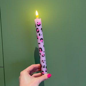 Op de afbeelding: Een brandende, lavendelkleurige kaars met een zwart en roze luipaardprintontwerp. De kaars wordt in een hand gehouden tegen een groene achtergrond. De vlam van de kaars is geel en oranje.