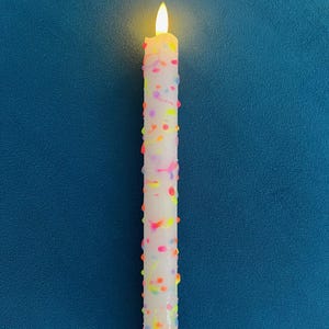 May include: A white, battery-powered candle with a flickering yellow flame. The candle is decorated with colourful, raised dots and speckles. The base of the candle is white with vertical ridges. The background is a solid, dark blue.