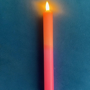 Op de afbeelding: Een lange, op batterijen werkende kaars met een kleurovergang, die overgaat van een helder roze basis naar een warm oranje aan de bovenkant. De kaars heeft een witte basis en een flikkerende, gele LED-vlam, wat een realistisch effect creëert.