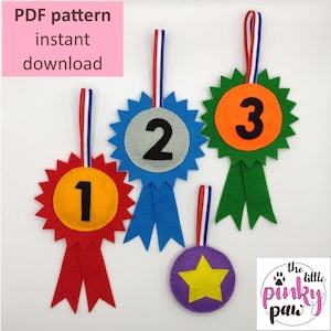 Könnte beinhalten: Ein Satz aus drei Filz-Preisbändern mit den Nummern 1, 2 und 3 darauf. Die Bänder sind rot, blau und grün. Es gibt auch einen lila Filzstern mit einem gelben Stern in der Mitte. Der Text "the little pinky paw" ist in der rechten unteren Ecke des Bildes zu sehen.