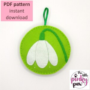 Könnte beinhalten: PDF-Muster für einen Filz-Schneeglockchen-Anhänger. Der Anhänger ist grün mit einer weißen Schneeglockchen-Blume. Das Muster ist zum sofortigen Download verfügbar.