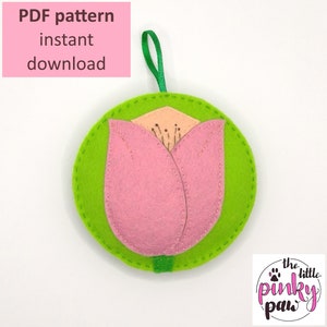 May include: PDF pattern for a felt tulip ornament. The tulip is pink with a green stem and leaves. The background is white with a repeating paw print pattern.