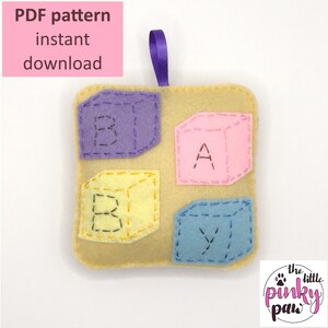 May include: A felt baby toy with four colorful blocks that spell out "BABY". The blocks are attached to a beige felt square with a purple ribbon.