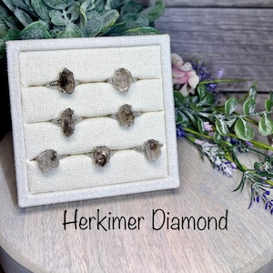 May include: A white display box with six rings featuring Herkimer diamond stones. The rings are arranged in two rows of three. The box is sitting on a wooden surface with the text "Herkimer Diamond" below it.